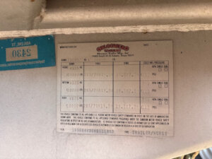 A metal trailer label displays information for a Bloomer Trailer, including VIN, tire sizes, rim sizes, and cold tire pressure. A blue label with the number 2490 is partially visible beside it. -Stillwater Trailer Sales