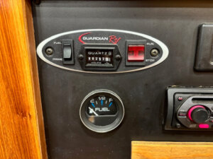 Close-up of an RV control panel showing a Guardian RV switch, an hour meter, a fuel gauge reading just above half, and part of an audio system. The panel is set in a wooden dashboard. -Stillwater Trailer Sales