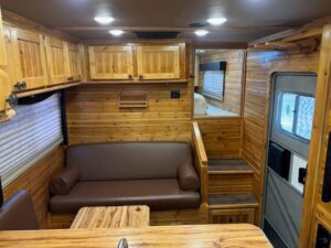 Interior of a camper van with wood-paneled walls and ceiling, brown cushioned sofa, overhead cabinets, a small bed area, and a door with a window; warm lighting creates a cozy atmosphere. -Stillwater Trailer Sales