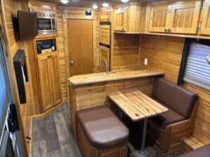 Interior of a small RV or camper with wooden walls, brown cushioned seating around a wooden table, a compact kitchenette with a sink, and cabinets for storage. The floor has a dark wood pattern. -Stillwater Trailer Sales