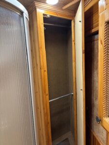 A wooden closet with an open door revealing an empty interior and a single metal hanging rod, next to a frosted glass shower stall in a rustic bathroom setting. -Stillwater Trailer Sales