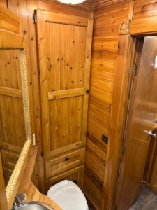 A small bathroom with wooden walls and cabinets, a built-in mirror, a sink in the corner, and a white toilet below the cabinet. The decor features rustic, natural wood finishes throughout. -Stillwater Trailer Sales
