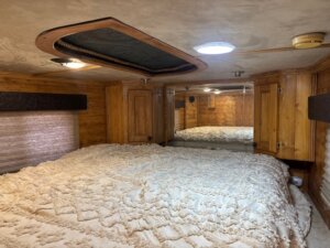 A cozy bedroom with wood-paneled walls and ceiling, a large bed with a textured white comforter, a ceiling hatch, round light, smoke detector, blinds, mirrors, and built-in wooden cabinets. -Stillwater Trailer Sales