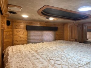 A cozy bedroom with wood-paneled walls, a large bed covered in a textured white bedspread, a window with a closed shade, and warm ceiling lights illuminating the space. -Stillwater Trailer Sales