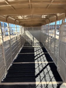 Interior view of an empty Swift Built 28' stock trailer with metal bars and a rubber mat floor, sunlight casting shadows inside, and buildings visible outside through the open end. -Stillwater Trailer Sales