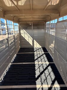 Interior view of an empty Swift Built stock trailer with metal walls, open windows on the sides, and a textured black rubber mat floor. Sunlight casts shadows through the bars onto the spacious 28' trailer floor. -Stillwater Trailer Sales