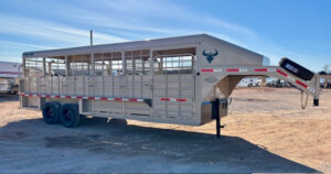 A beige gooseneck livestock trailer with dual axles, metal rails, and a bull head logo is parked on a dirt lot under a clear blue sky. -Stillwater Trailer Sales