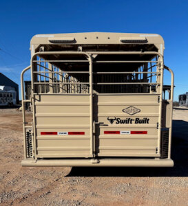 The rear view of a tan livestock trailer with metal bars and the brand name &ldquo;Swift-Built&rdquo; visible. The trailer is parked on gravel with a clear blue sky in the background. -Stillwater Trailer Sales