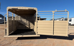 A tan livestock trailer with its rear gate open stands on gravel under a clear blue sky, ready for loading or unloading animals. -Stillwater Trailer Sales