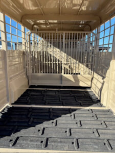 Interior view of an empty tan livestock trailer with metal bars, gridded windows, and a rubber mat floor, taken in daylight. Sunlight casts shadows inside the trailer. -Stillwater Trailer Sales
