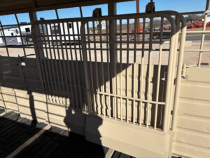 Two beige metal gates with vertical bars inside a livestock trailer, with sunlight casting shadows. Outside, vehicles and equipment are visible through the trailer&rsquo;s open slats. -Stillwater Trailer Sales
