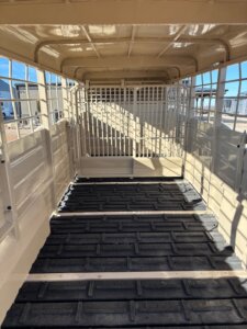 Interior view of an empty, light-colored livestock trailer with metal bars and gates, black non-slip floor, and sunlight casting shadows inside. Industrial buildings and blue sky are visible through the trailer windows. -Stillwater Trailer Sales