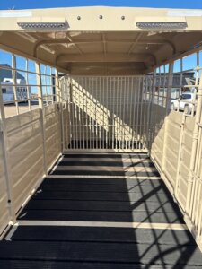 Interior view of an empty beige livestock trailer with metal bars, an open back, and sunlight casting shadows on the floor. Buildings and a white vehicle are visible in the background. -Stillwater Trailer Sales