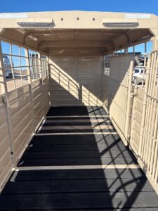 Open, empty tan livestock trailer with metal sides and roof, black non-slip floor, and open rear gate. Sunlight casts shadows inside. Vehicles and buildings are visible in the background. -Stillwater Trailer Sales