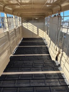The interior of an empty livestock trailer with beige metal walls and black rubber flooring, featuring sunlight streaming through the side openings and casting shadows. -Stillwater Trailer Sales