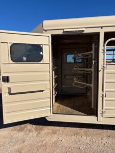 A tan metal horse trailer with its side door open, revealing a wooden floor and three empty saddle racks mounted inside against the interior wall. The trailer sits on dirt under a clear blue sky. -Stillwater Trailer Sales