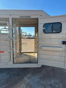 Open door of a beige trailer revealing an empty interior with racks, likely for saddles, and a view of a dirt lot and blue sky in the background. -Stillwater Trailer Sales
