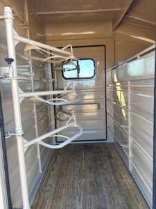 Interior of a horse trailer featuring a wooden floor, saddle racks on the left wall, metal slats along the right wall, and a closed door with a window at the far end. -Stillwater Trailer Sales