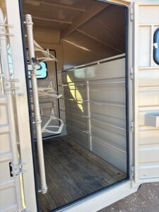 Open door of an empty beige livestock or horse trailer, showing interior with wood floor, metal walls, and saddle racks attached to the left side. Sunlight partially illuminates the inside. -Stillwater Trailer Sales