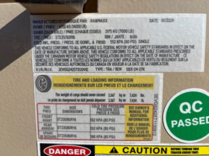 Close-up of vehicle information stickers showing tire and loading data, weight specifications, manufacturing details, and warnings, with a green QC Passed sticker partially visible on the right side. -Stillwater Trailer Sales