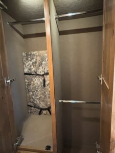 An empty wooden closet with open doors, double hanging rods, and a marbled panel secured with black straps inside on the left side, designed for the 2019 Bloomer 5 Horse trailer. -Stillwater Trailer Sales