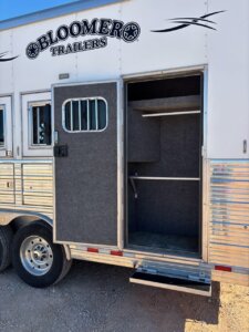 The open side door of this 2019 Bloomer 5 Horse trailer reveals a carpeted interior with shelves, a hanging rod, and a fold-out step leading up from the gravel outside. -Stillwater Trailer Sales