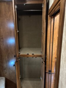 Tall, empty wooden cabinet with two compartments, separated by a single shelf in the middle, and open doors revealing the interior. The cabinet is positioned next to a wooden wall and door. -Stillwater Trailer Sales