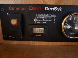 Close-up of a Cummins Onan GenSet control panel showing a digital hour meter reading 586.9 hours, indicator instructions, a backlit switch, and part of an analog gauge on a wooden surface. -Stillwater Trailer Sales