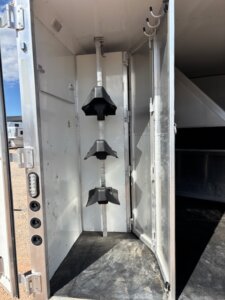 The image shows the open rear door of a horse trailer, revealing a vertical saddle rack with three black saddle holders inside a clean, metal compartment. Hooks are attached to the upper right wall. The trailer is parked on dirt. -Stillwater Trailer Sales