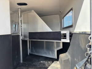 Interior of a horse trailer with metal dividers, padded barriers, a grated window on the right, and a non-slip black floor; the area is clean and well-lit with white walls and ceiling. -Stillwater Trailer Sales