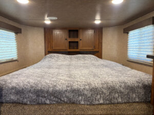 A cozy bedroom with a large bed covered in a gray blanket, wooden cabinets at the head of the bed, beige walls, and two windows with closed blinds on either side, softly lit by ceiling lights. -Stillwater Trailer Sales