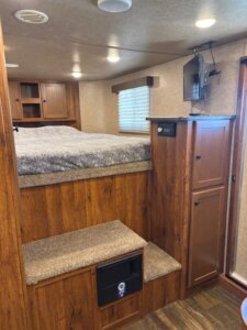 Cozy RV interior with a raised bed, wooden steps leading up to it, built-in cabinets, a small wall-mounted TV, window with blinds, and wood flooring. The space appears compact and efficiently designed. -Stillwater Trailer Sales