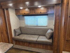 A brown cushioned sofa with two matching pillows sits below a window with blinds, framed by wood-paneled walls and cabinets in a cozy RV interior. -Stillwater Trailer Sales