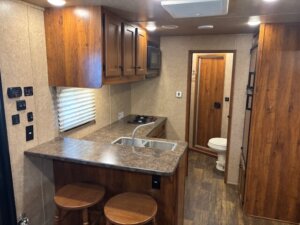 A compact RV kitchen with wood cabinets, a sink, stove, microwave, and counter with two wooden stools. In the background, there is a small bathroom with a toilet and wooden flooring throughout the space. -Stillwater Trailer Sales