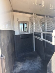 Interior of a clean, empty horse trailer with padded dividers, black flooring and walls, metal bars, and a small window near the front. -Stillwater Trailer Sales