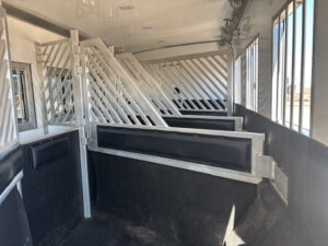 Interior of an empty horse trailer with metal dividers and padded walls, designed to safely transport multiple horses in separate stalls. Sunlight is streaming through the barred windows. -Stillwater Trailer Sales