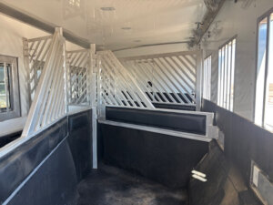 The interior of a horse trailer with metal dividers and padded sides, creating individual stalls for transporting horses. Sunlight streams through windows on the right. -Stillwater Trailer Sales