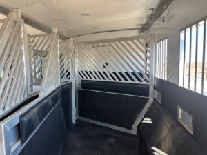 Interior of a livestock or horse trailer with metal dividers, rubber flooring, and barred windows; sunlight is shining through the windows on the right side. -Stillwater Trailer Sales