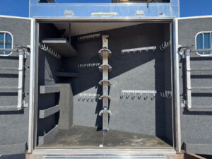 Open storage compartment with gray carpeted walls, multiple hooks and racks on the sides and back, and vertical saddle racks in the center, designed for organizing horse tack and equipment. -Stillwater Trailer Sales