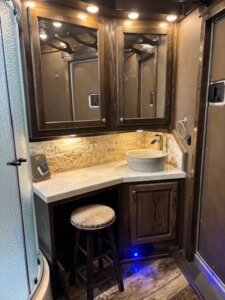 A rustic bathroom with dark wood cabinets, a round stone vessel sink on a beige countertop, a decorative stool, a glass shower door, and warm under-cabinet lighting. -Stillwater Trailer Sales