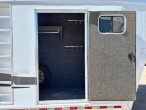 Open door of a trailer reveals a compartment with hooks on the wall, a shelf, and a spare tire inside. The exterior is metallic with a small window on the door. -Stillwater Trailer Sales