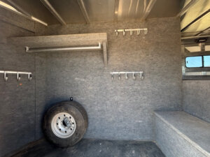 Interior of a gray, carpeted utility vehicle with metal shelves, several hooks on the walls, a spare tire leaning against the wall, and a small bench-style platform on the right side. -Stillwater Trailer Sales