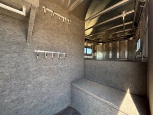 Interior of a horse trailer showing a gray carpeted tack room with metal hooks on the walls, a bench, windows, and an upper storage area with metal framing. Sunlight comes in through the side window. -Stillwater Trailer Sales
