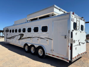 A large white horse trailer with six windows, triple axles, and decorative black graphics is parked on a gravel lot under a clear blue sky. -Stillwater Trailer Sales