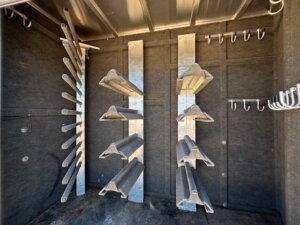Interior of a horse trailer tack room with multiple angled saddle racks and bridle hooks mounted on carpeted walls. The metal racks and hooks are empty, and sunlight filters through the space. -Stillwater Trailer Sales