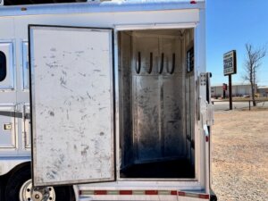 The image shows an open compartment on a metal trailer with three saddle racks inside. The trailer is parked on gravel near a road, with a Trailer Sales sign visible in the background. -Stillwater Trailer Sales