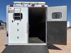 Open rear doors of a white horse trailer with a ramp lowered to the ground, revealing a dark, empty interior with dividers. The trailer is parked on a dirt lot under a clear blue sky. -Stillwater Trailer Sales