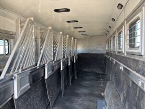 Interior of an empty livestock trailer with individual metal stalls, barred windows on the right, and a clean, textured black floor and walls. The space is well-lit from windows and overhead lights. -Stillwater Trailer Sales