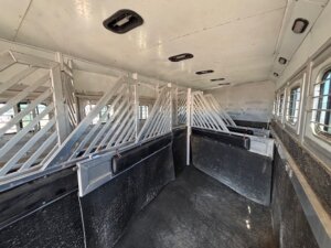Interior of a horse trailer with empty stalls separated by metal dividers, barred windows on the right side, and overhead lights on the ceiling. The floor and walls are lined with protective material. -Stillwater Trailer Sales