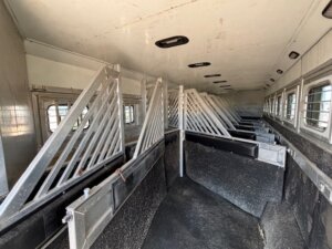 The image shows the interior of a livestock trailer with metal dividers, rubberized flooring, and windows along the sides for ventilation. The stalls are empty and clean, ready for transporting animals. -Stillwater Trailer Sales